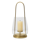 A brass lantern-style Nilo candle holder from Broste Copenhagen with a clear glass shade and a single cream pillar candle inside, shown on a plain white background.