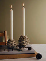 A dining table scene with a set of two lit ceramic sculptural candle holders shaped like pine cones featuring a glossy off-white glaze with subtle brown speckling and warm tan edges