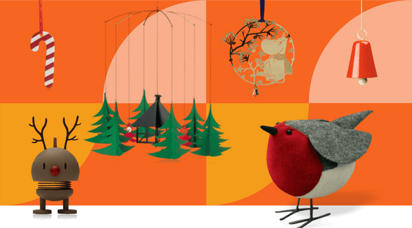 Joyful Scandinavian Christmas decorations including striped candy cane ornament, Flensted Christmas trees mobile, Moomin ornament, orange metal bell, Hoptimist bouncing reindeer figure and large red felt robin on bright orange background