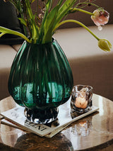 Green glass vase by Broste Copenhagen with flowers on a side  table with candle