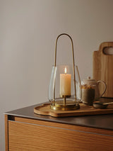 A brass lantern-style Nilo candle holder from Broste Copenhagen with a clear glass shade and a single cream pillar candle inside, shown on a sideboard with wooden tray