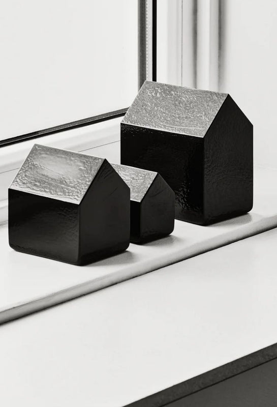 Three small black house-shaped decorative glass objects by Kosta Boda are arranged on a white windowsill in natural light.