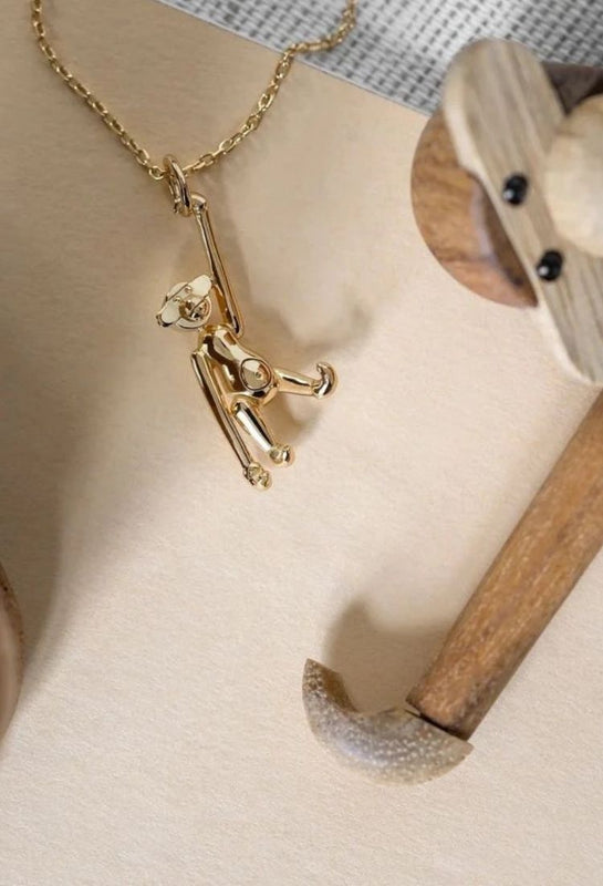 A gold-plated monkey necklace based on Kay Bojesen's Danish wooden monkey, with a glimpse of the original iconic design in the background