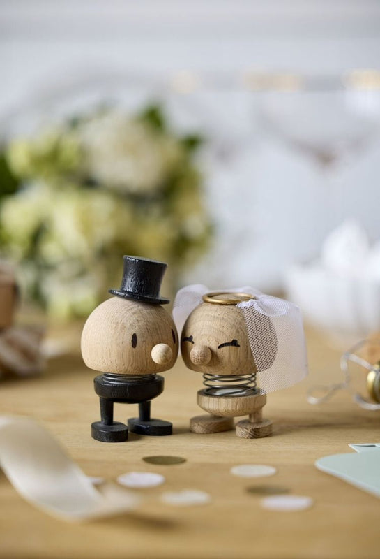 Two small oak Hoptimist wedding figurines on a wooden tabletop: one a briedgroom with a black top hat and black body, the other a bride with a white veil. The background is softly blurred with flowers.