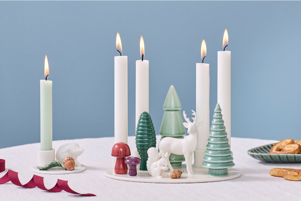 Dottir Woodland Stories ceramic advent candle holder, with squirrel, deer, trees and mushrooms, with lit candles on a Christmas-themed table on a blue background