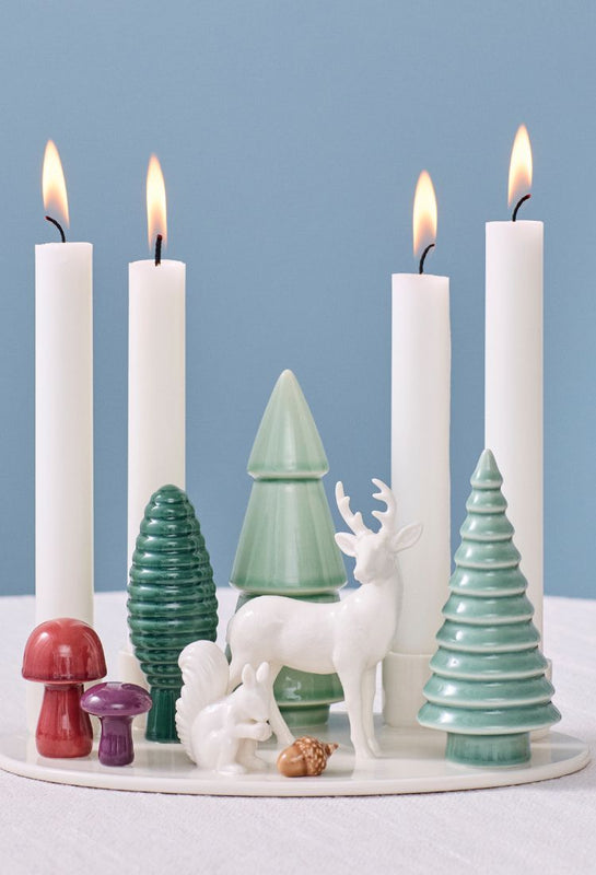 Deatil of Dottir Woodland Stories ceramic advent candle holder, with squirrel, deer, trees and mushrooms, with lit candles on a Christmas-themed table on a blue background