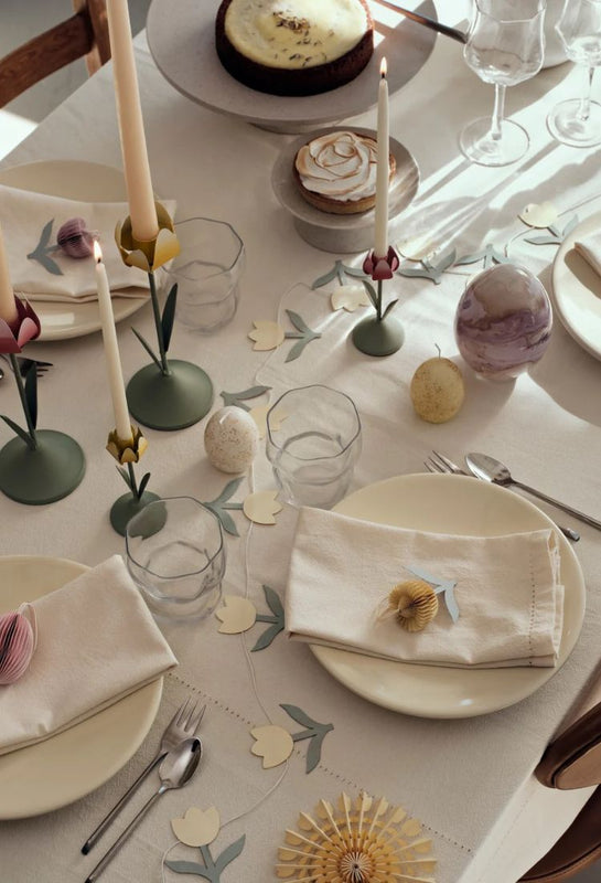 Easter-themed table setting using products by Broste Copenhagen with candles and decorations including eggs, and flowers.