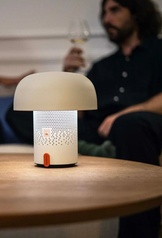 White Kooduu sensa play mini speaker lamp on a wooden table, person holding wine glass in the background