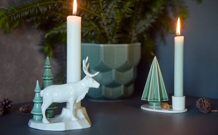 A festive holiday table setting featuring a white ceramic Dottir Nordic Design reindeer candle holder and a Chritsmas tree candle holder, both with white candles, styled with pinecones and a potted plant in a green Dottir pot in the background.