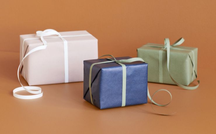 Three wrapped gift boxes on a warm tan background: a large pale blush box with a white ribbon, a medium blue box with a pale green ribbon, and a medium sage green box with a light green ribbon.