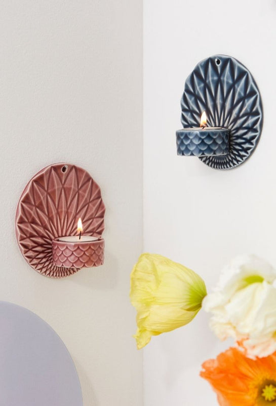 Two textured ceramic wall sconces by Dottir Nordic Design holding lit tea light candles, one in dusty pink and one in dark blue, mounted on a white wall with yellow and orange flowers in the foreground.